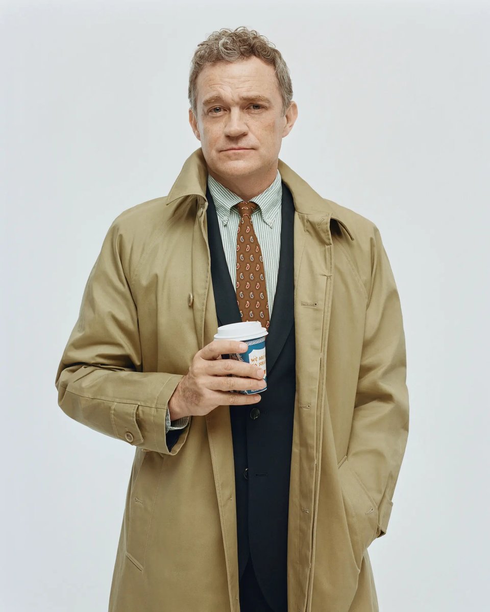 Patrick Radden Keefe, middle-aged white man with graying red hair, dressed in a trench coat, a blue striped button down, paisley red tie, holding a classic Greek style New York coffee cup. White background.