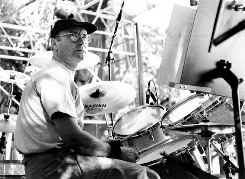 A photo of the drummer Phil Collins, sitting a drum kit, looking upwards and to the right. He is onstage outdoors, we can see the edges of a stage behind him. The photo is black and white and sunny