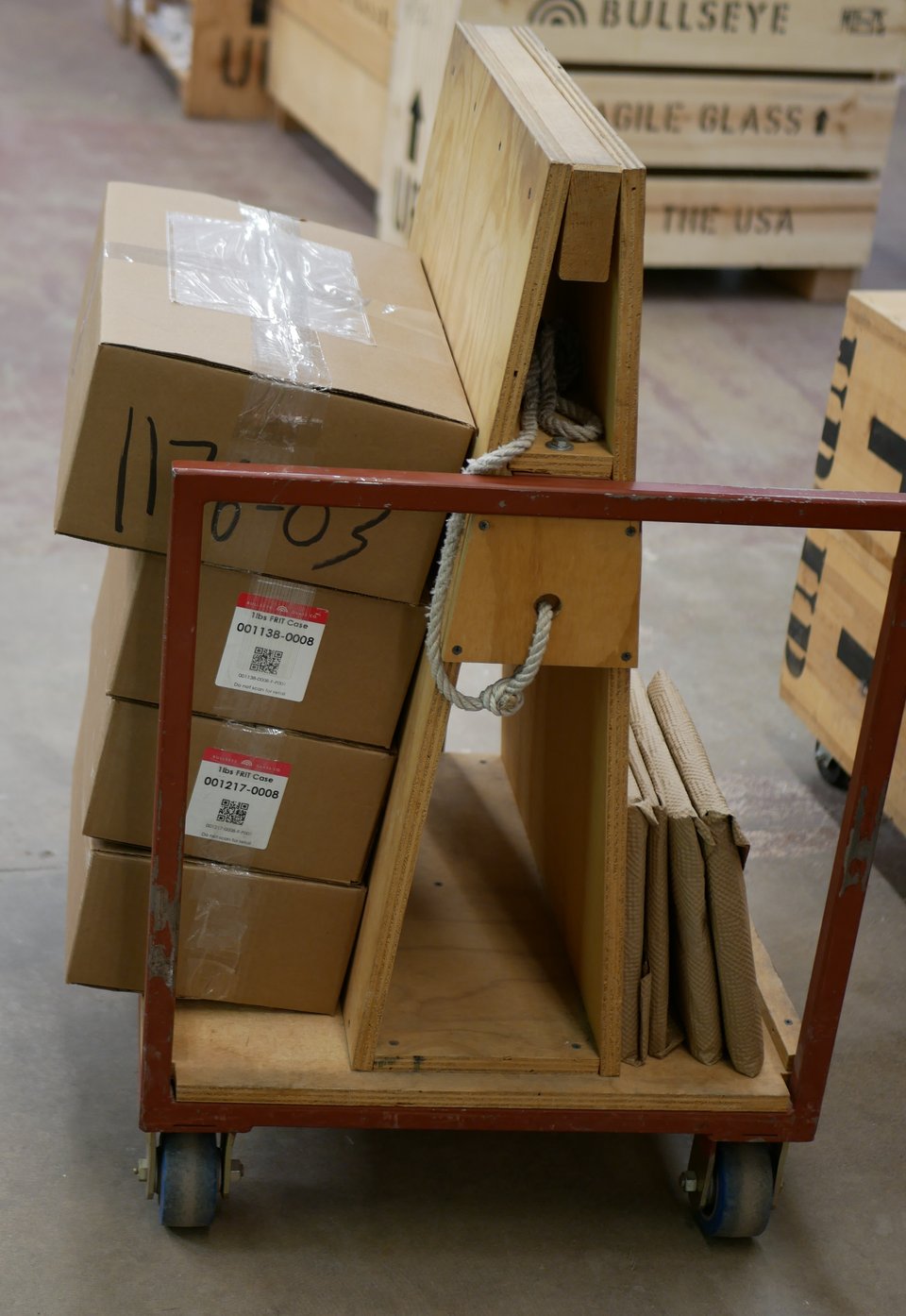 a flatbed metal cart with a plywood A-frame in the middle for leaning panes of glass on. one side has a bunch of packages of 10 inch glass squares, wrapped in paper, the other side contains piles of boxes