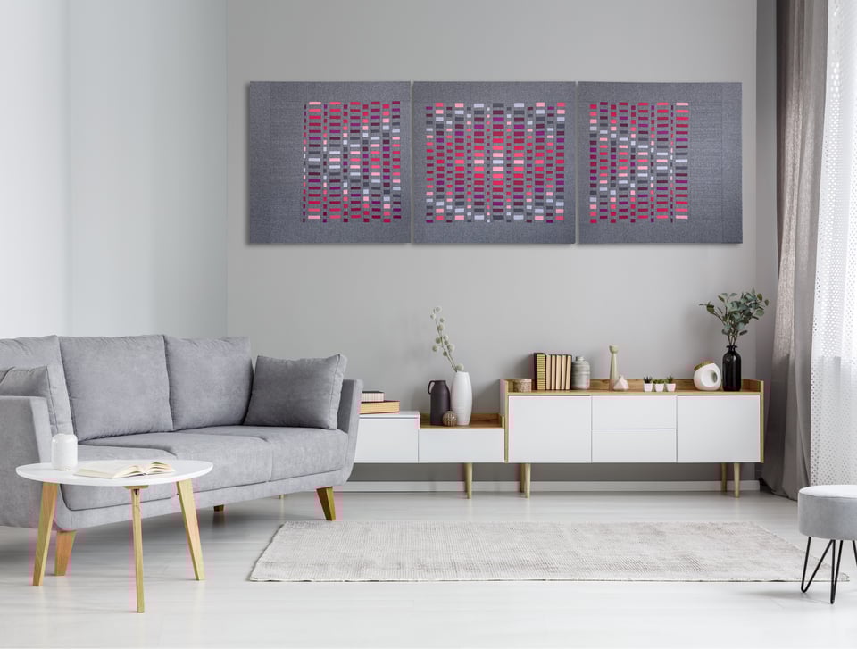 A big and colorful, geometric artwork made with textiles is hanging on the wall of a living room. The scene is calm and peaceful.