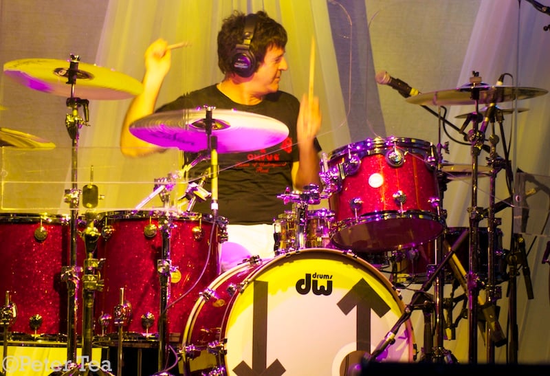 A photo of the drummer Clem Burke playing the drums. His arms are up in the air, holding to drum sticks, and his head is tilted off to the right. He is wearing headphones. The drum kit is red, and he is bathed in a yellowish light.