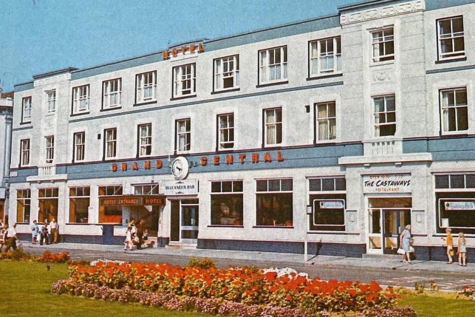 A photo of the Grand Central in the 1960s. It has three storeys. The ground floor is large plate windows, and the upper floors are sash windows. within the front there are two sections that jut out slightly and have decorative balconies and pediments. It's front has some blue detailing with lettering in red on it. There is a flowerbed full of red geraniums in the grass out front: the kind of municipal planting that is thankfully a thing of the past now.