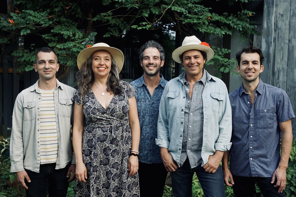 Four men and a woman in a patterned dress stand in a line in front of foliage.