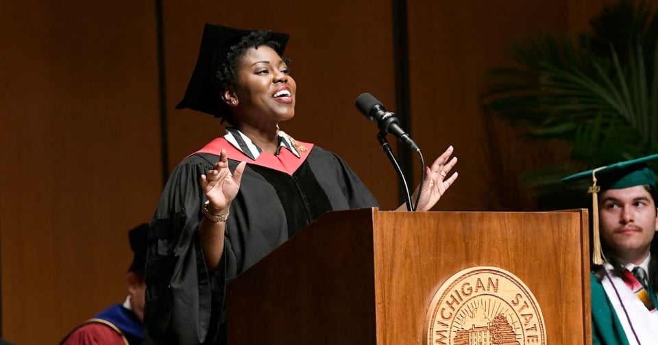 A commencement speaker in academic regalia stands at a podium, speaking into a microphone during what appears to be a graduation ceremony. The wooden podium features the engraved seal of Michigan State University, indicating the event's location.