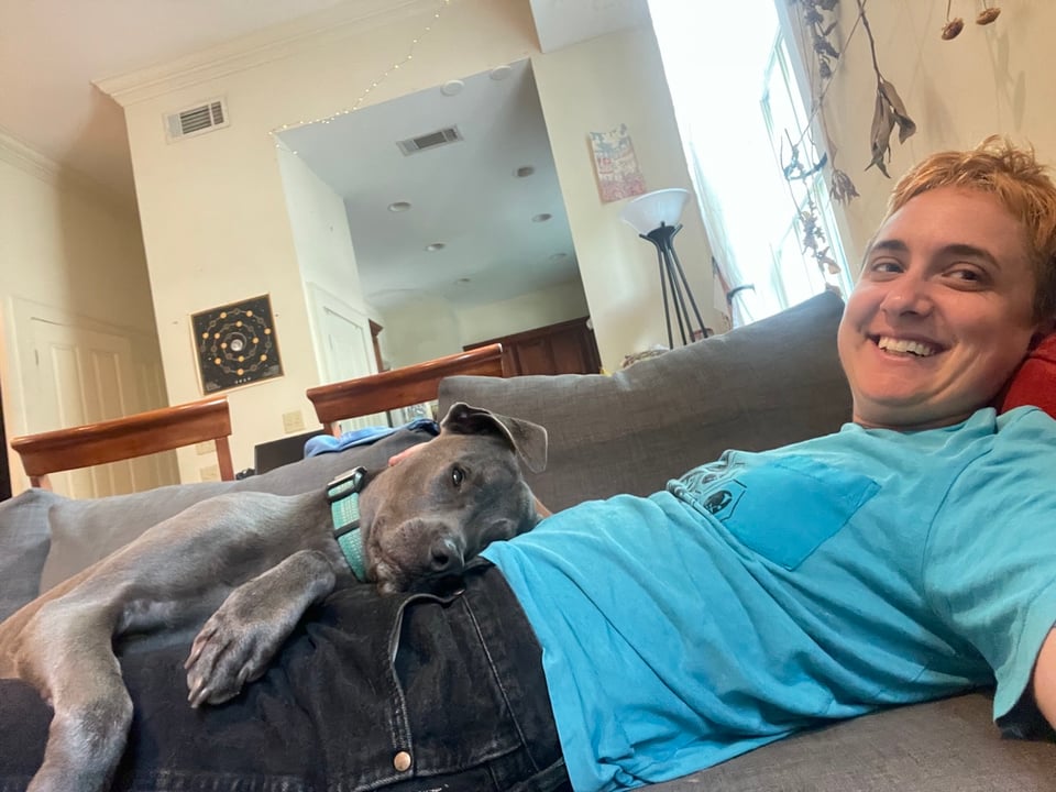 A grey floppy eared dog laying on a smiling white person with bleached hair and a blue tshirt. the dog is looking at the camera