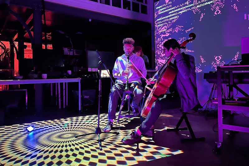 Two performers playing cello and clarinet, with computer generated projections on the floor and on the wall behind them.