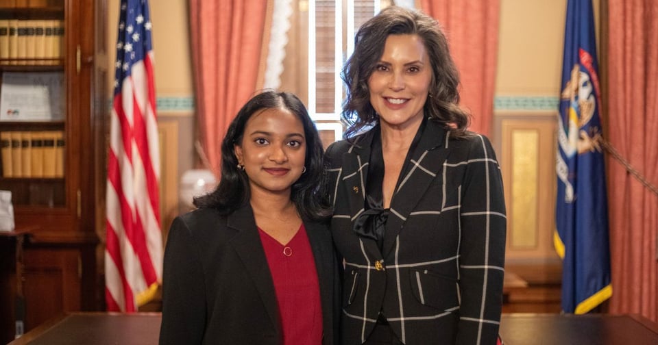 JMC student and Michigan Gov. Gretchen Whitmer stand side by side smiling at the camera. They are positioned indoors, with an American flag and another official-looking flag visible behind them, suggesting a formal or governmental setting.