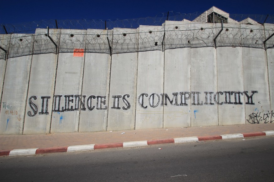 Tall concrete barriers topped with barbed wire on the side of a road. Stenciled on the wall are the words: SILENCE IS COMPLICITY