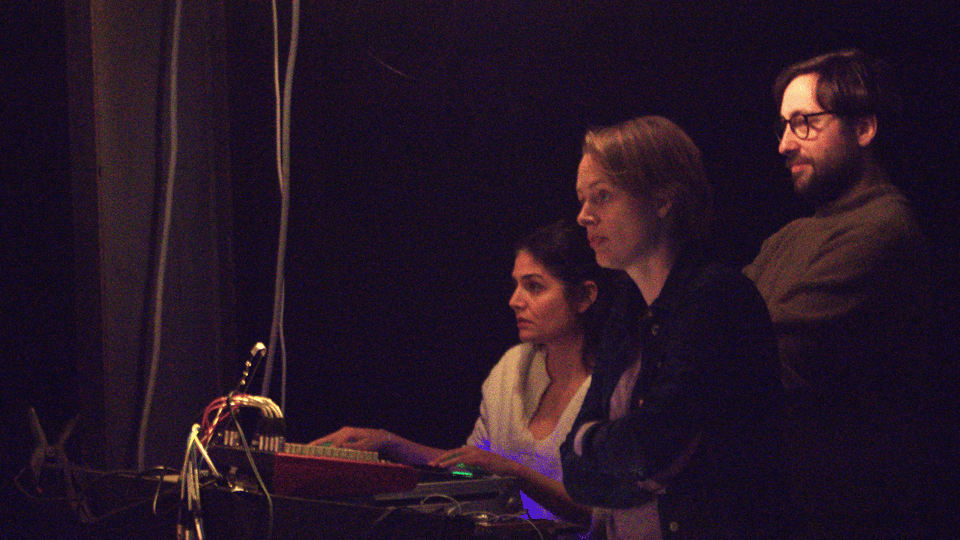 Three people (Tanine, Laura and Gael) behind a sound table and a light board, in the tech booth of a theatre