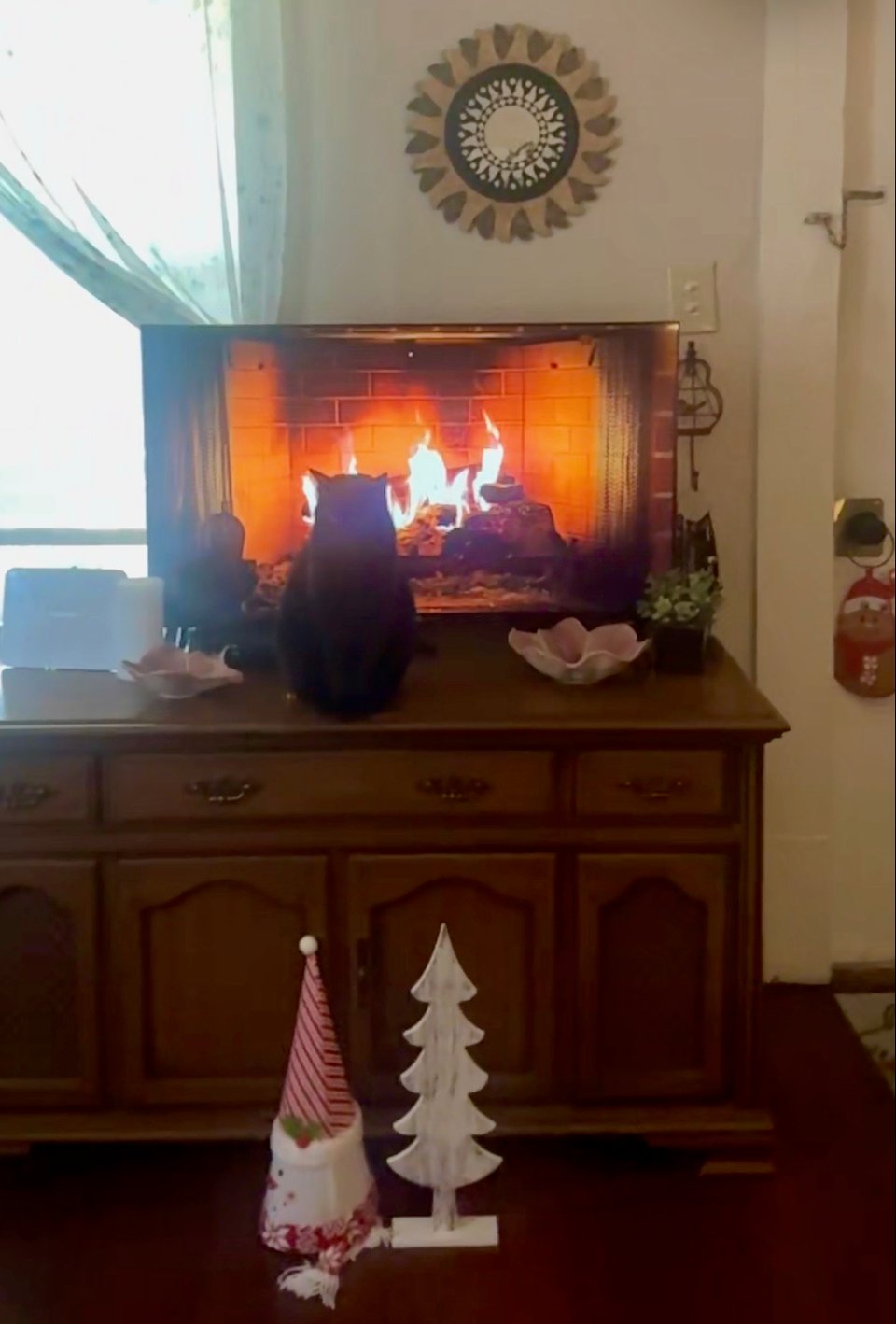 Morpheus, my sleek black cat, sitting on my cabinet-style record player in front of the TV, where a fireplace scene is playing. There are bright orange and yellow flames coming from stacked logs inside a red brick fireplace, and his back is to the screen, as though he's trying to warm himself. There are also decorations visible, including two pink flower-shaped decorative bowls, a small fake potted plant, a sunburst mirror on the wall, a Santa stocking hanging to the side, and on the floor in front of the record player, a white wooden Christmas tree and an upright red and white striped Christmas hat.