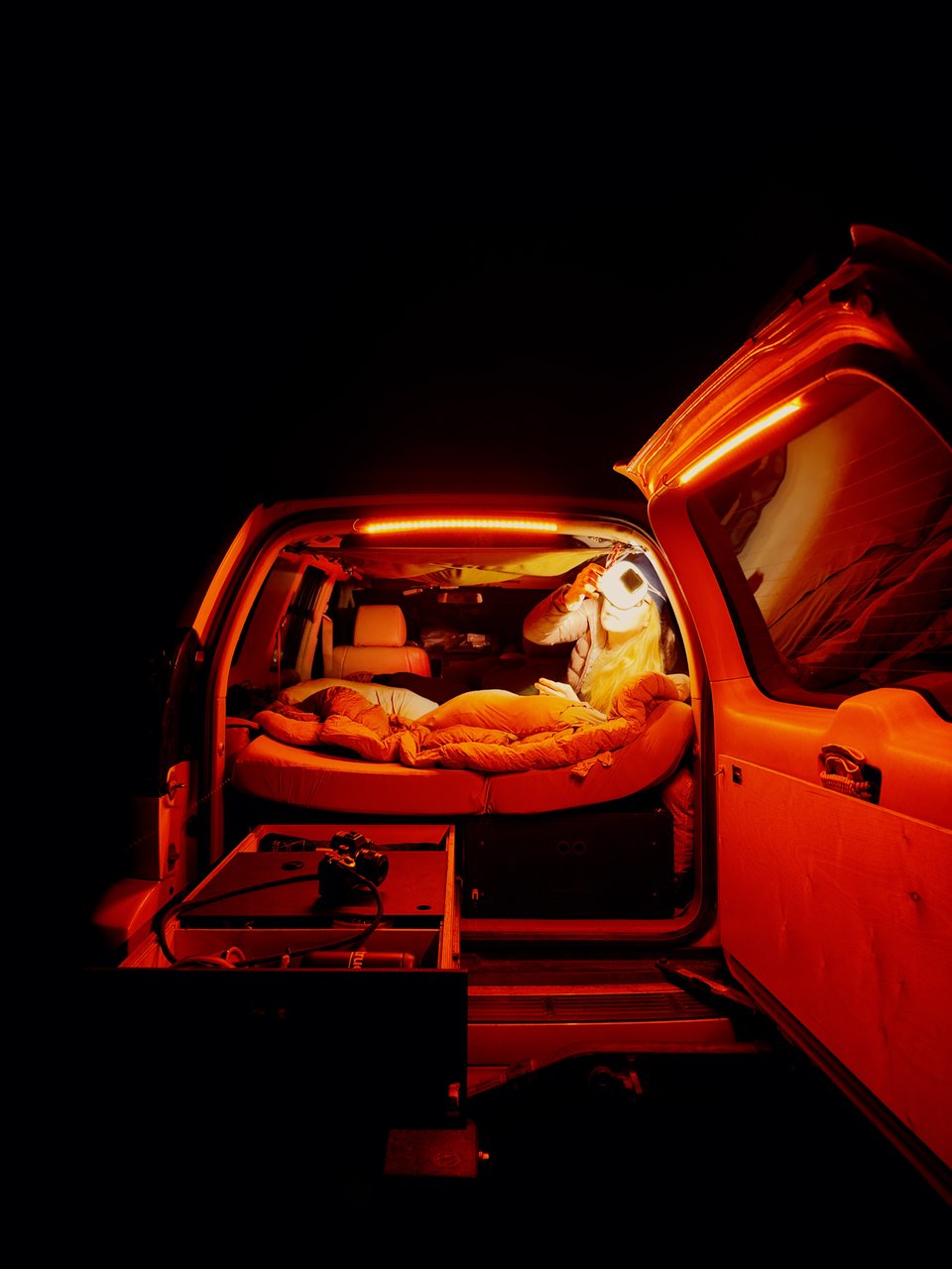 Jen setting up inside the back of our rig. I'm outside taking the image, bathed in red light as the barn door of our car is wide open, and one of the long drawers below the mattress is fully extended.