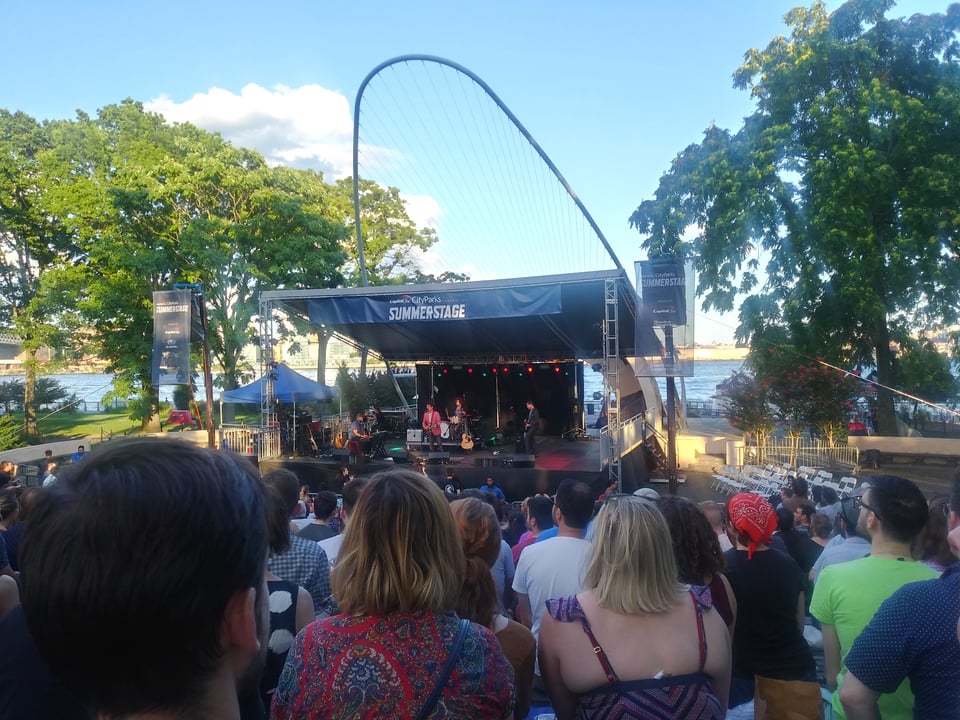The Mountain Goats performing on stage at an outdoor theater, a crowd looking on. Trees are on either side of the stage, and behind the stage is the East River.
