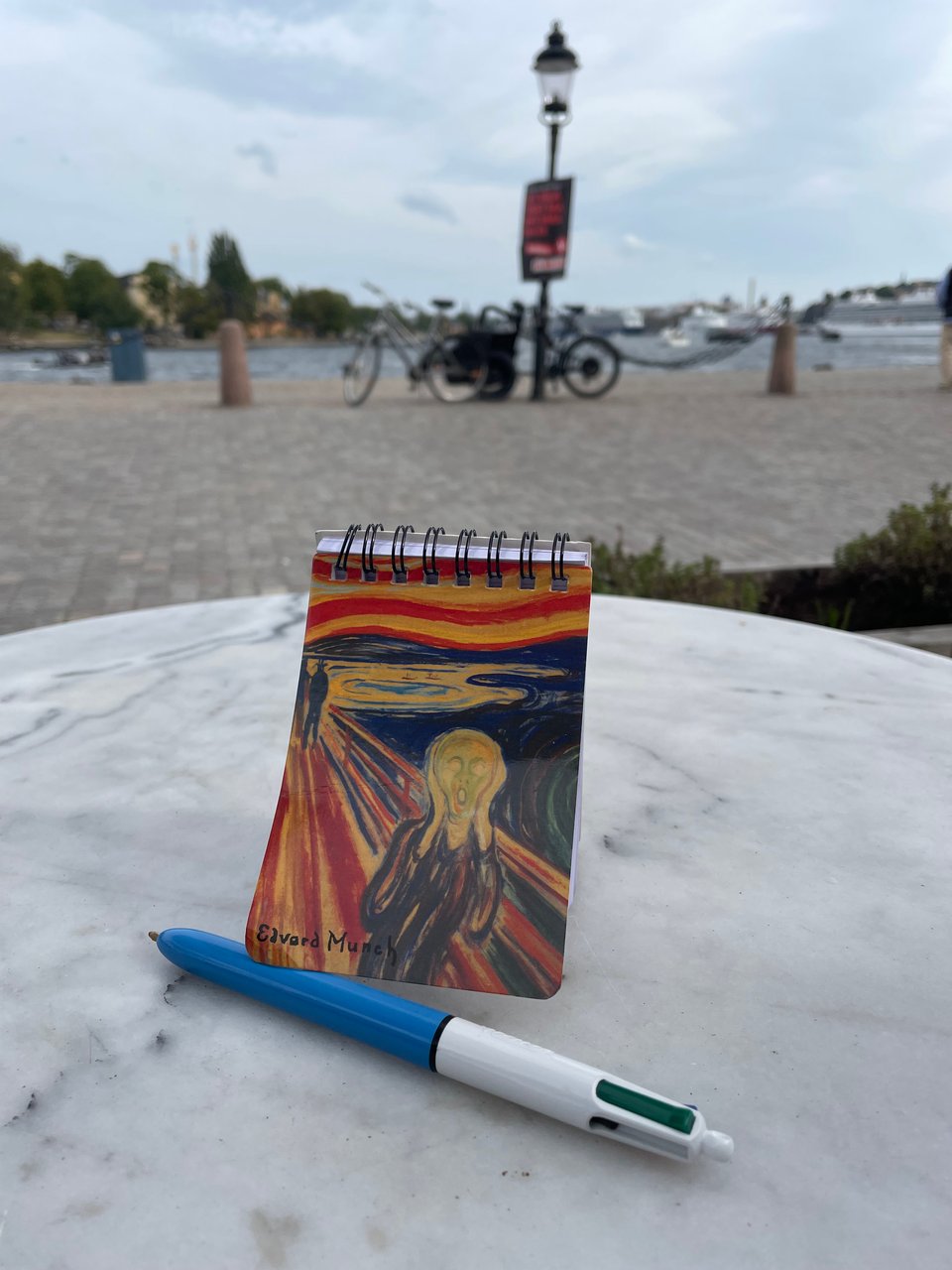 A notebook with Munch's Scream on it and a multi-color pen on a cafe table by open water.