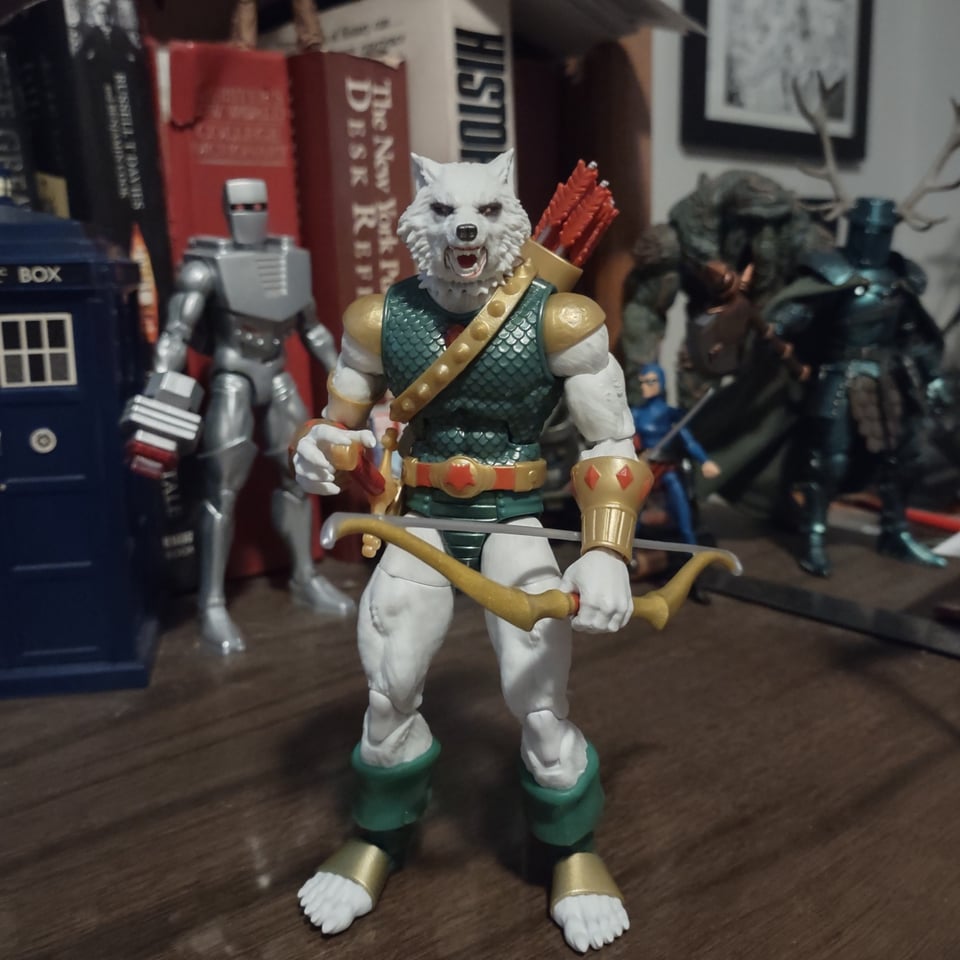 An action figure of a white-furred wolf man stands on a wooden desk with other figures behind him. He wears green and gold fantasy-esque armor, with a bow in one hand and a quiver slung over one shoulder. His other clawed hand rests on the pommel of a sword hanging on his belt.