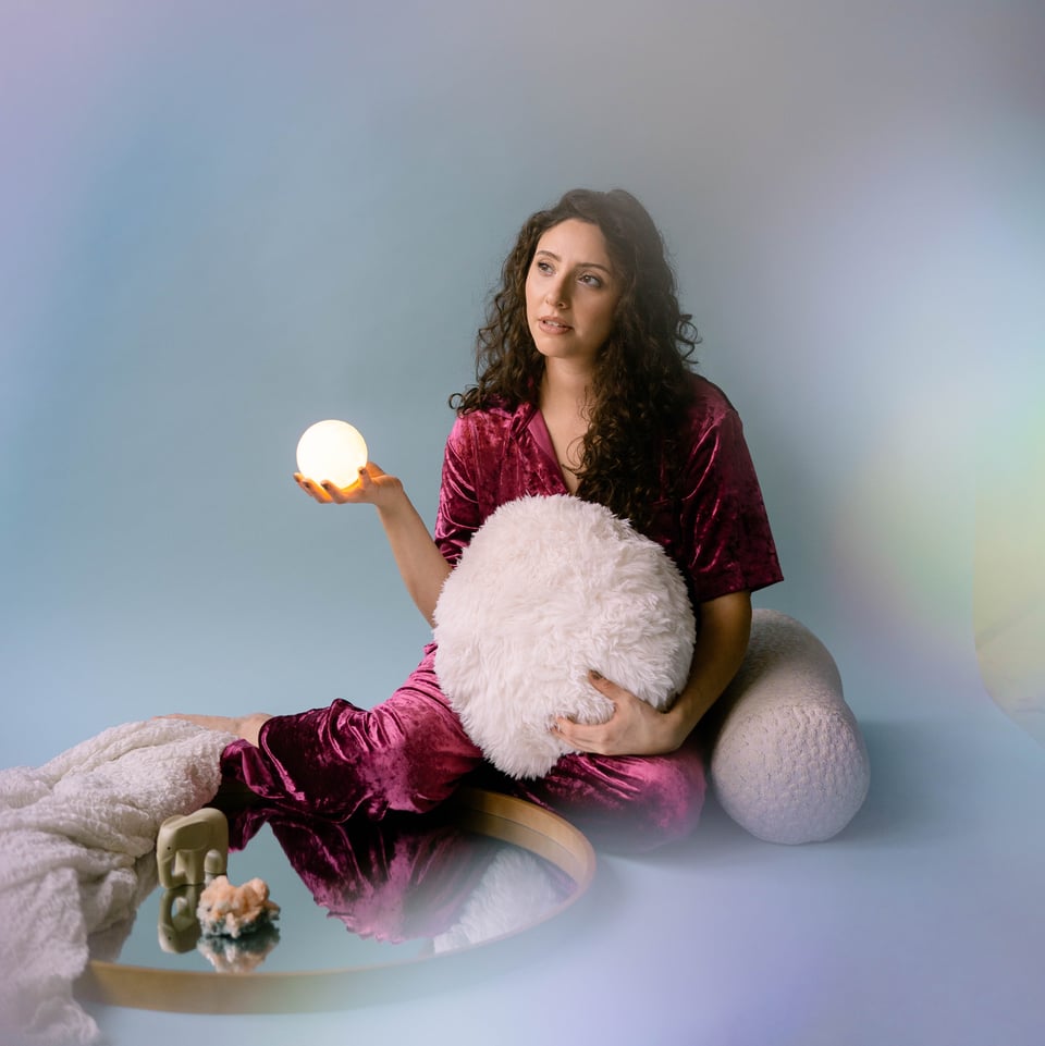 A photograph of Victoria Fragoso seated in a misty setting next to a mirror. She is wearing a purple robe and is holding a small glowing white orb and a large furry pillow.