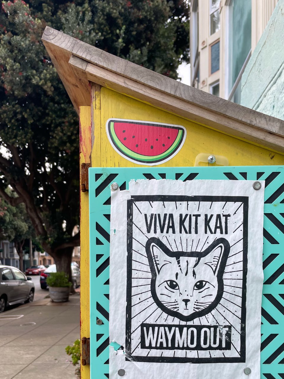 a Little Free Library in San Francisco with a watermelon sticker and a flyer with a cat on it that says, "Viva Kit Kat, Waymo Out"