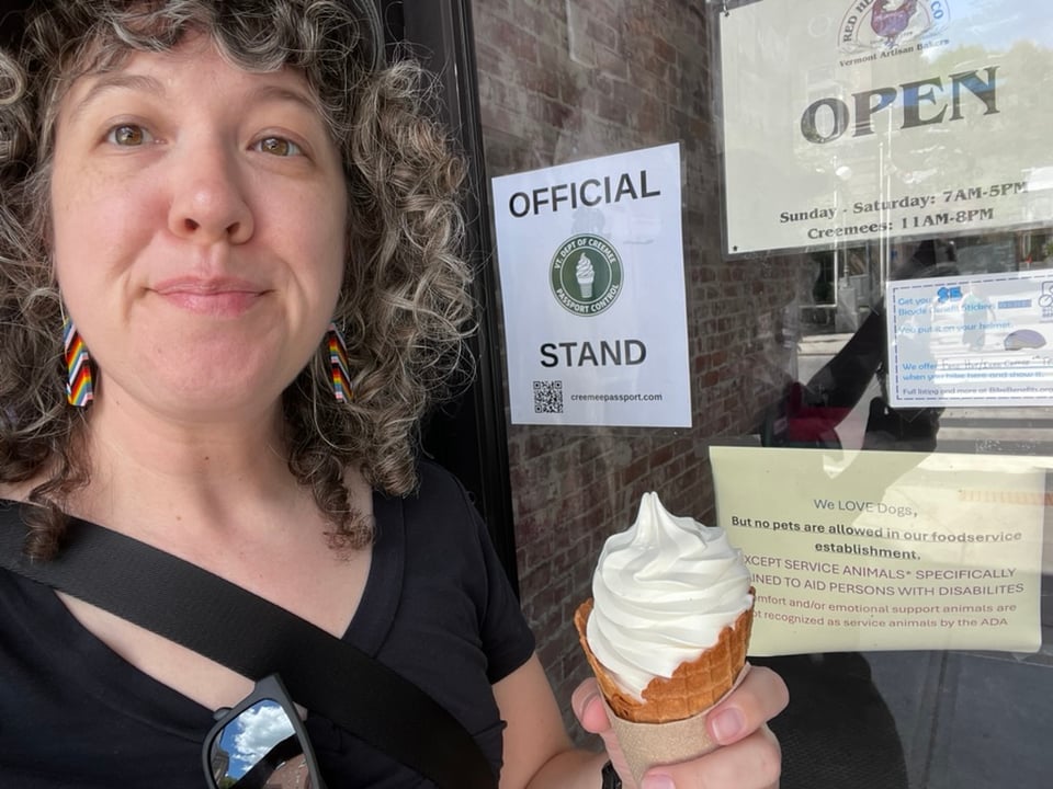 Photograph of Eira, a white woman with curly hair, dressed in a balck shirt and rainbow earrings, holding a maple creemee in front of a store.