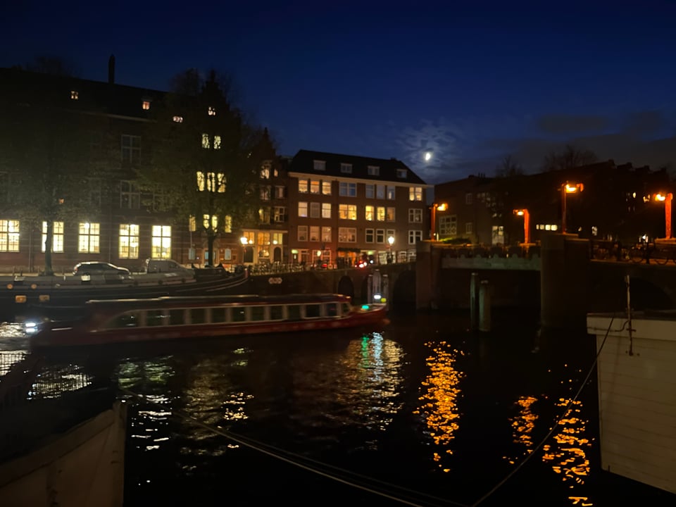 A nighttime view of an Amsterdam canal with the quarter moon rising over the city. The houses have lights on in the windows and a long low canal boat glides through the water.