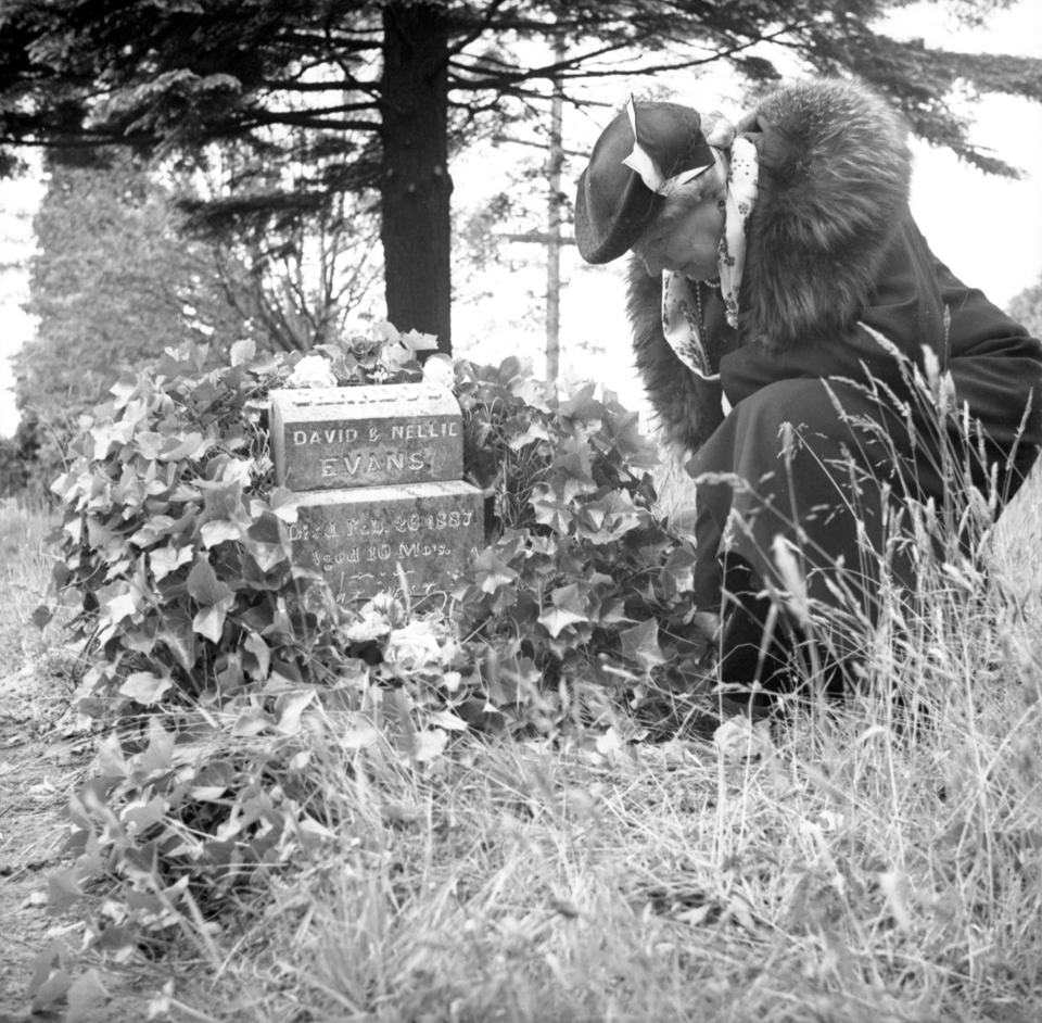 Image of a woman in a winter coat and hat kneeling next to a small stone grave marker where you can read "David and Nellie Evans" and beneath is a marker for Caradoc Evans, 10 months.