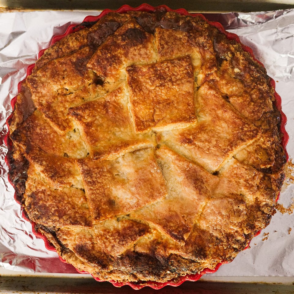A pie topped with squares of dough in a shingled pattern sits in a red pie pan on top of a sheet pan covered in tinfoil.