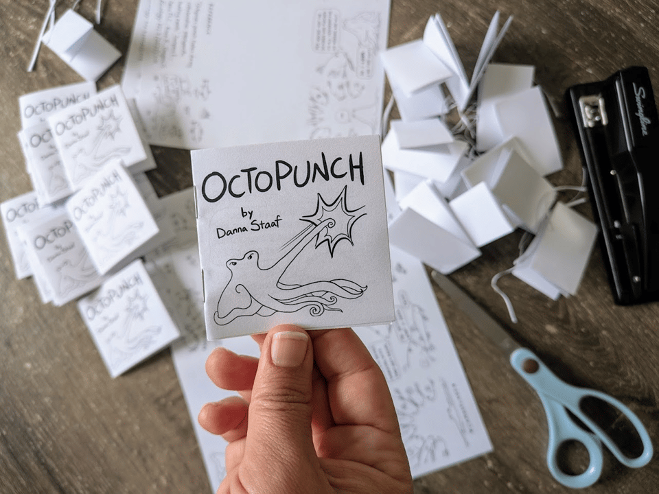 Photo of my hand holding a mini one-sheet-of-paper zine with all the zine-making materials on a table in the background: unfolded papers, scissors, stapler, scraps of paper and a pile of finished zines. The cover says "OCTOPUNCH by Danna Staaf" and has a drawing of an octopus giving a big comic book punch with one arm.