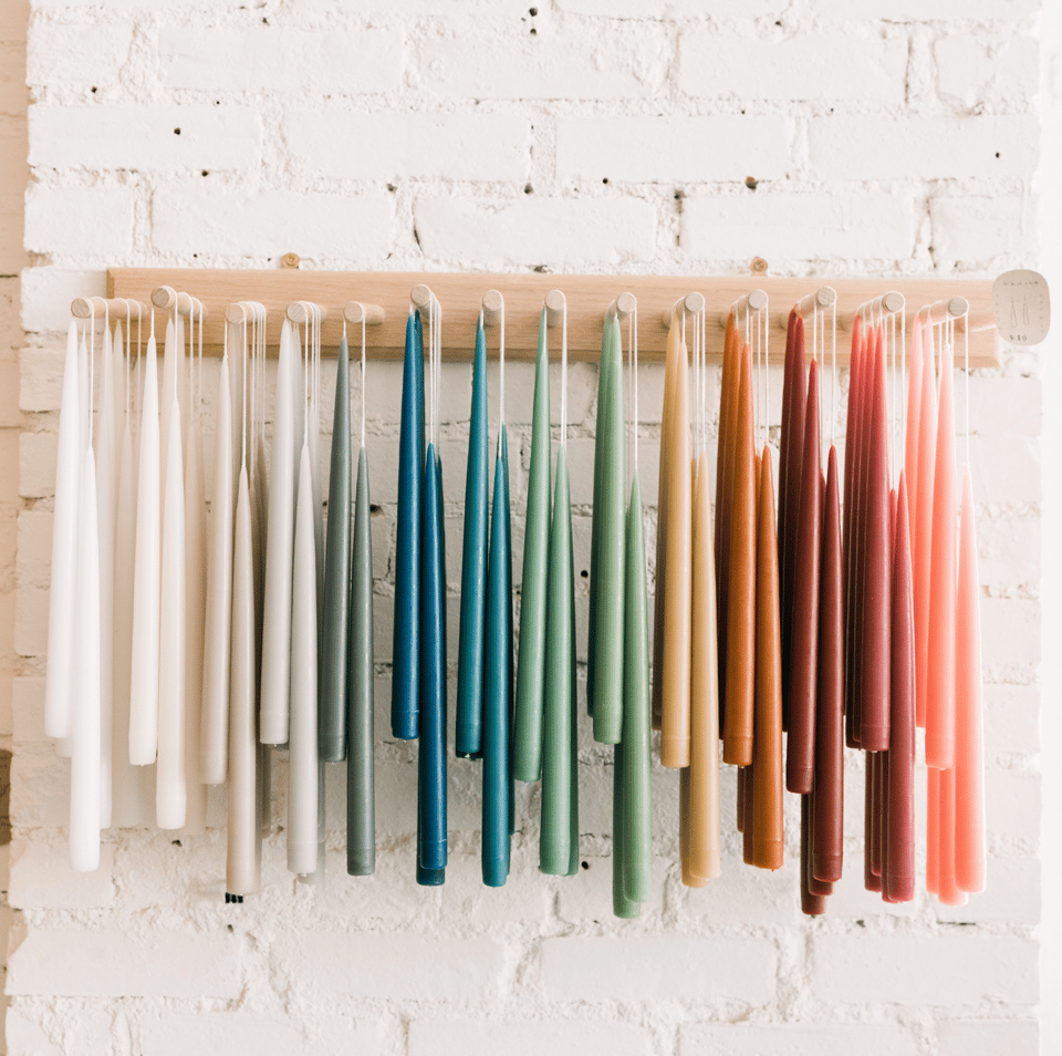 An array of beautifully colored beeswax candles handing from a wooden shelf against a white wall.