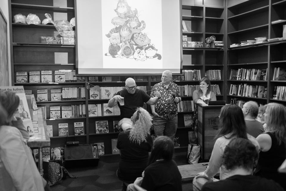 Photo in a bookstore with a projection screen with audience backs in foreground left and right and three people (Brian Basset, Glenn Fleishman, Georgia Dunn) at front of room, with Brian gesturing and Glenn and Georgia looking at him.