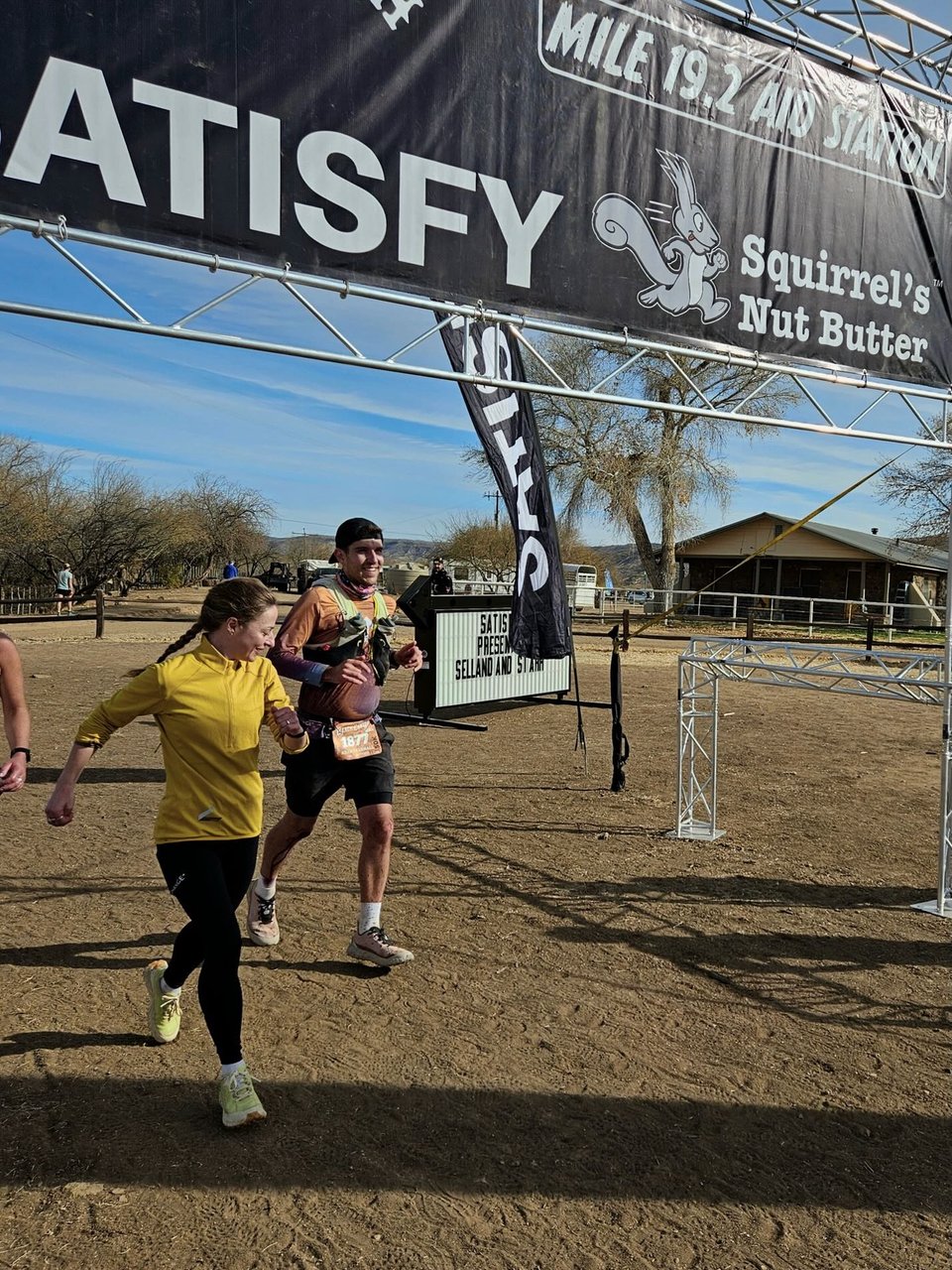 Kelsey, wearing a yellow quarter-zip, black tights, and green-ish running shoes next to Adam wearing an orange and purple shirt, running underneath a black banner at Bumble Bee aid station.