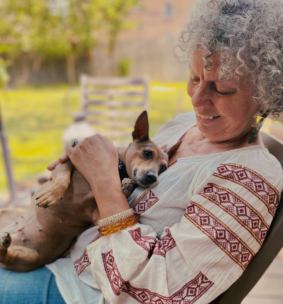 Woman with curly gray hair smiles gently while cradling a relaxed small dog. She wears a patterned blouse and bracelets. A warm, serene outdoor setting.