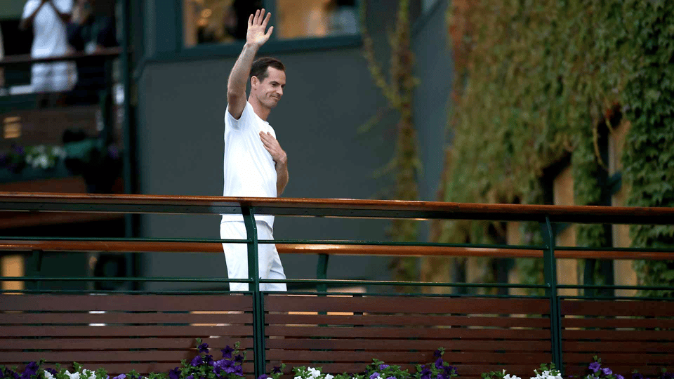 Andy Murray, one hand on his chest and one raised, waves to the crowd (out of frame) from the Wmibledon bridge off of Centre Court, the court and its ivy in te background