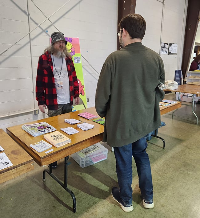 A guy is standing in front of a table, talking to another guy who is behind the table.