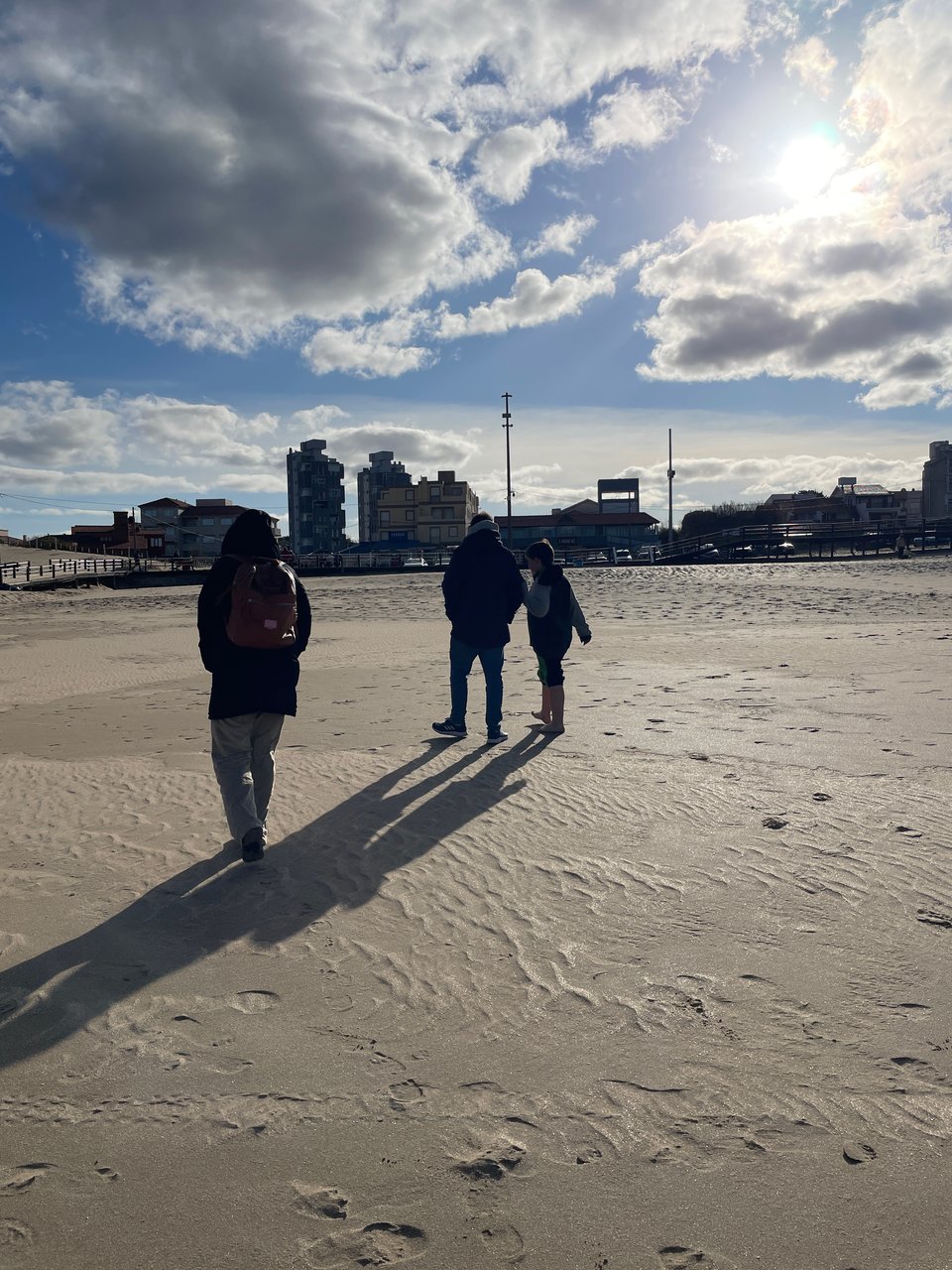 Three people bundled up in jackets walk on the beach away from the camera. Their long shadows are cast on the sand in the afternoon sun. The blue sky is filled with white puffy clouds pushed around by the wind.