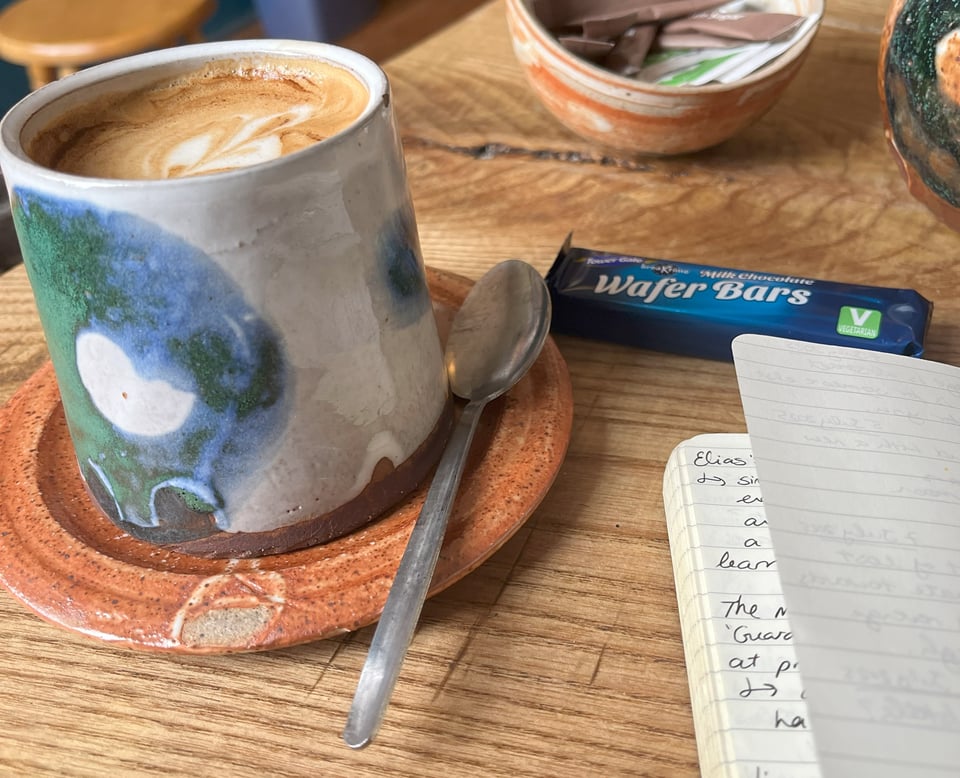 A photograph of a coffee mug on a worn wooden table. The mug is squat and cylindrical, with abstract shapes in blue and green glaze. The saucer it sits on looks like a rusty, textured surface. A metal teaspoon rests on the saucer. In the bottom right corner, a lined notebook page can be seen, though the words are illegible. A wafer bar in a blue packet rests can be seen in the background.
