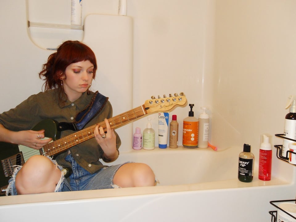 A woman with her reddish hair pulled back wearing a button-down shirt and ripped jeans with her knees poking out sits in a bathtub playing a Fender Telecaster guitar.