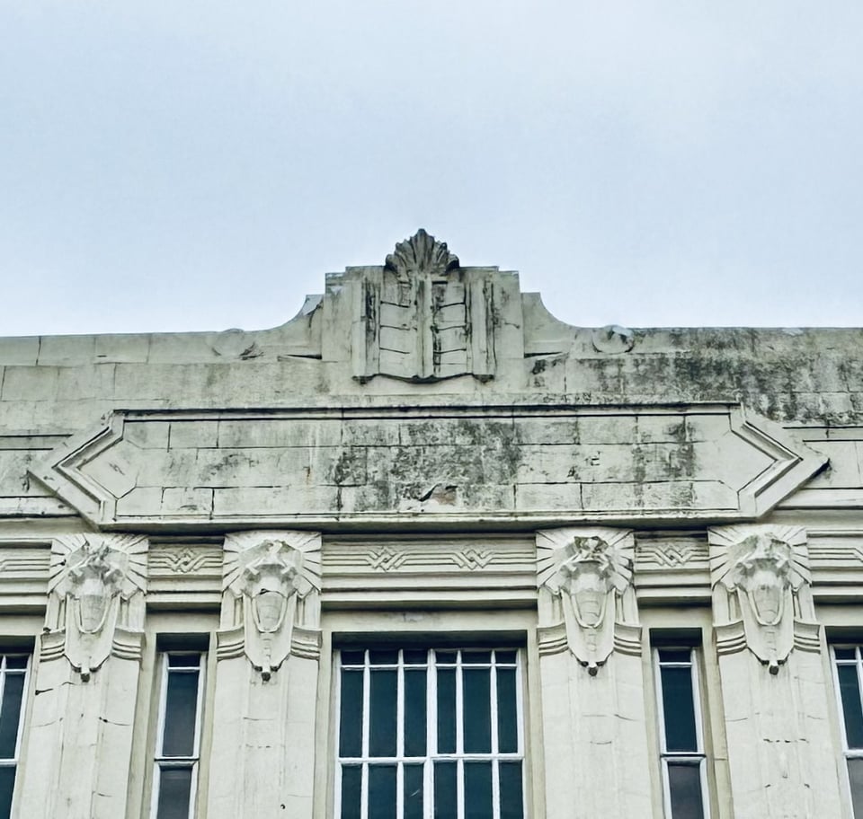 Close up of the top of the building. It's of stone and has a pediment with an open book on it. Four of the columns between the windows are visible, and the capitals are revealed as having stylised elephant heads on them.