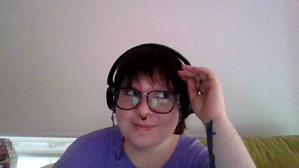 Ren is a white non-binary person with dark hair, wearing black glasses, and black headphones. They are smiling and staring right off-screen.