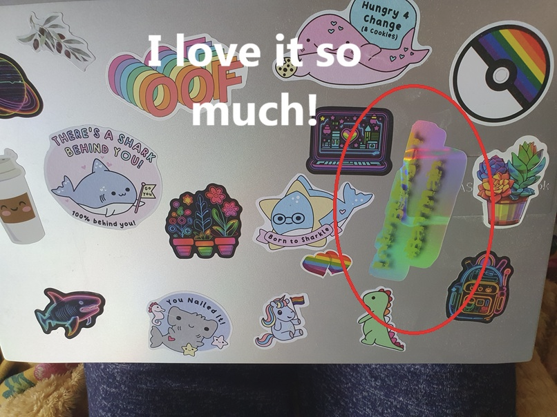 Photo of the back of my laptop, covered in various fun and rainbowy stickers with a new holographic one circled, that says 'Low Budget #Batman'. Text overlaid says 'I love it so much!'