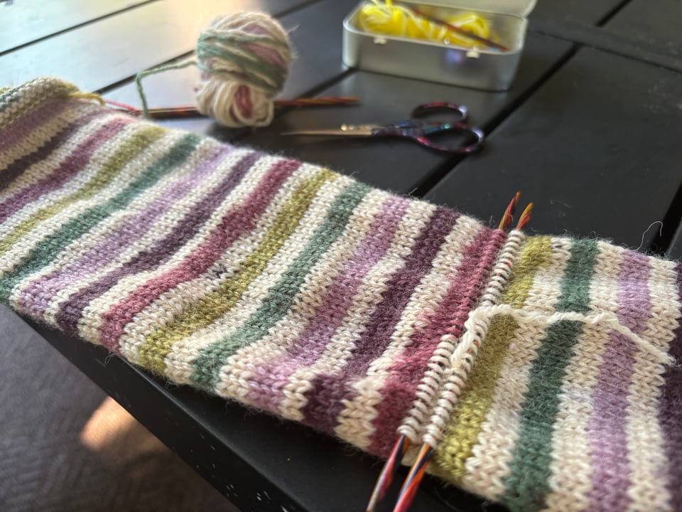 Close up of a multicolour knitted tube with two rows of stitches midway down picked up on wooden double-pointed needles; the row between them has been snipped and the yarn partially pulled out to make room for an afterthought heel.