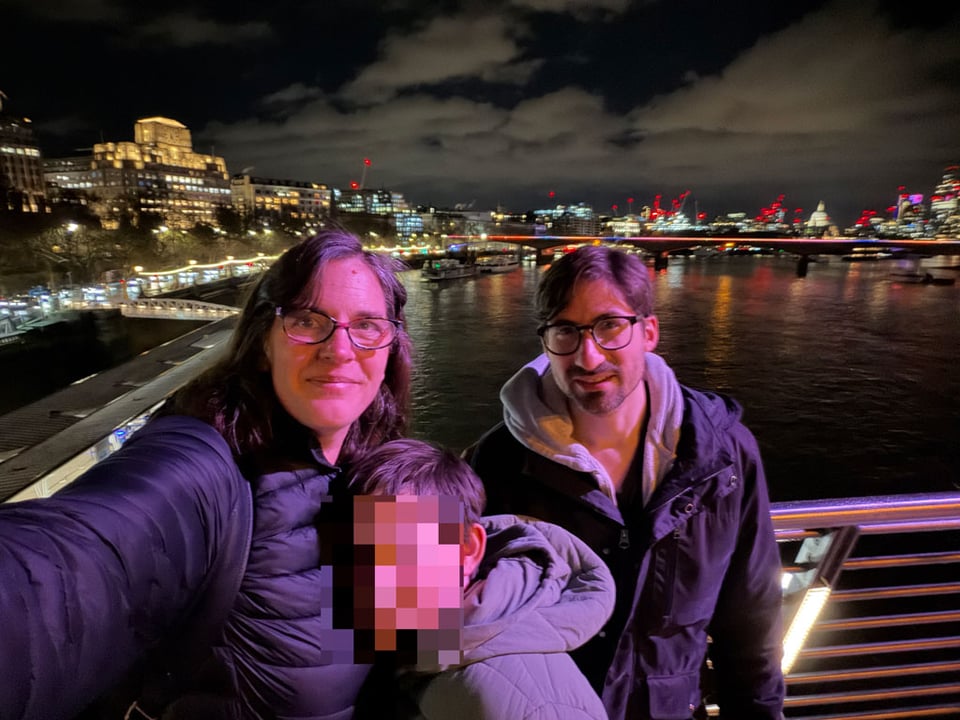 A woman, man and their child smile at the camera on a bridge over the London Thames. The child's face is blurred out.