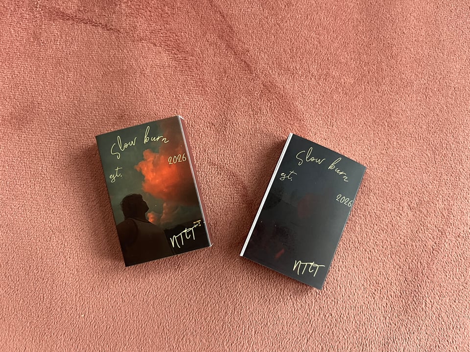 Two Slow burn matchboxes against rose gold velvet. One shows the front album art, a picture of Nola in shadow looking up at vivid pink-gold clouds against a blue-grey sky and the other shows a very dark picture of Nola with a guitar in deep shadow and dim campfire light. Both have light yellow cursive text that says "Slow burn," "est. 2026," and "NTtT."