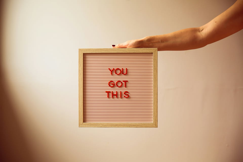 Image of an off-white background and a message board with the words, "You got this" in capital red letters on the board. A hand is rested on top of the board.