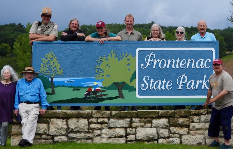Usually, when you drive into a Minnesota state park, you’re welcomed by a sturdy brown sign with gold lettering. We’re good with that—nothing wrong with those dignified workhorses. But … Frontenac State Park is special! We’ve long had a beautiful, colorful painted entrance sign, but last year, it keeled over from wear and tear. Behold—we have a brand new one! Some of the FSPA members who helped our park purchase this unique sign recently posed for the photo above, clambering all over it like giddy kids as they cracked ridiculous jokes. From left, they are: Kristen Eide-Tollefson, Ted Tollefson, park manager Jake Gaster, Janet Malotky, Pamela Miller, Dean Potter, Rachael O’Donnell, Susan Wandmacher, Wayne Wandmacher and Steve Dietz, who snapped this photograph and then raced back to the sign so he’d be in the photo too. Thanks to all of you and the rest of FSPA for making our special sign possible!