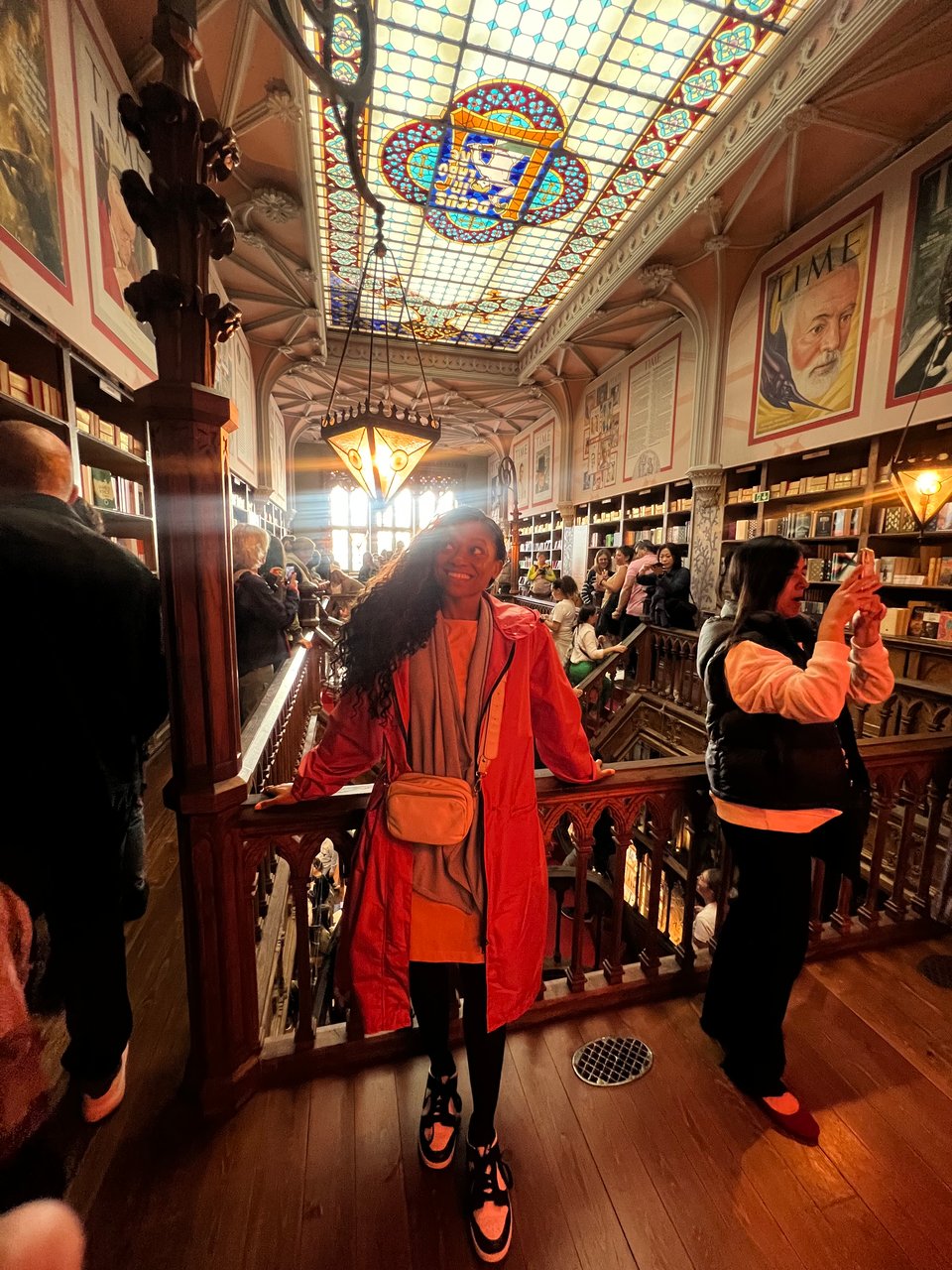 I am standing in Livraria Lello, an historic bookstore, dressed in a long, pink trench coat, smiling, and looking up and away. The interior features ornate woodwork, stained glass ceilings, and walls adorned with portraits and literary decorations, evoking a warm, classic atmosphere.
