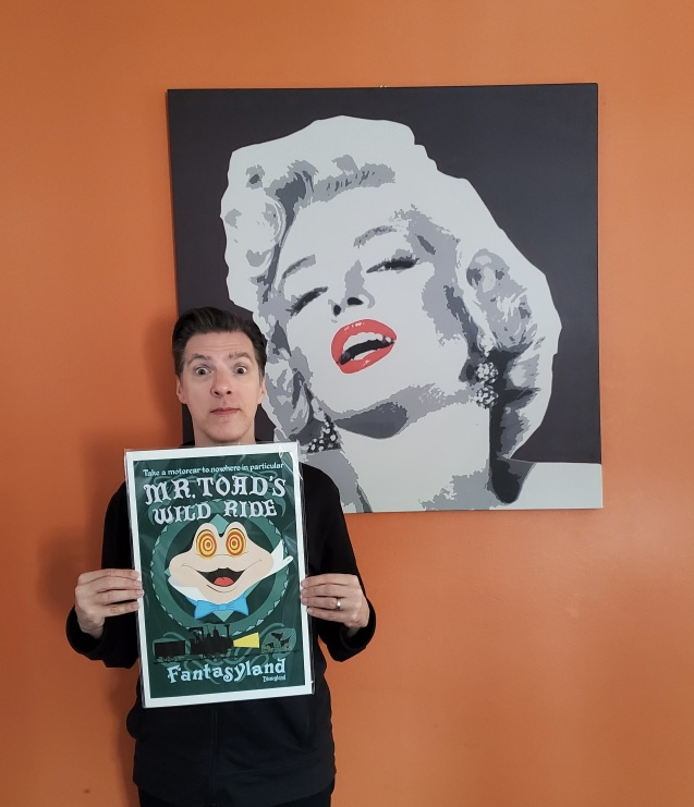 A man holds a Mr Toad's Wild Ride poster in front of a Marilyn Monroe painting