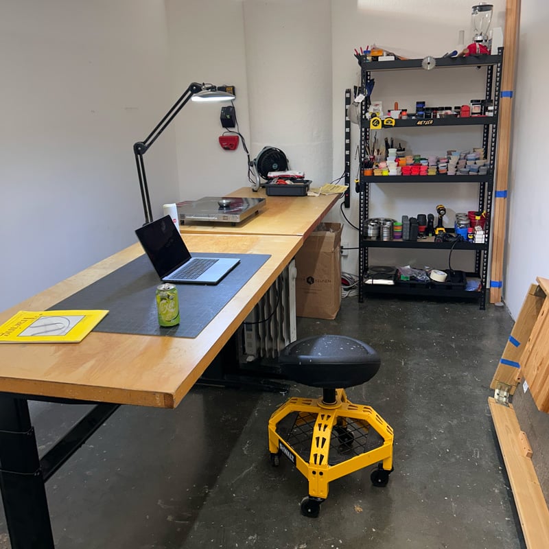 A recently set up and relatively clean art studio. There's a couple of tables, a rolling stool, and a shelving unit along the back wall filled with assorted crap.
