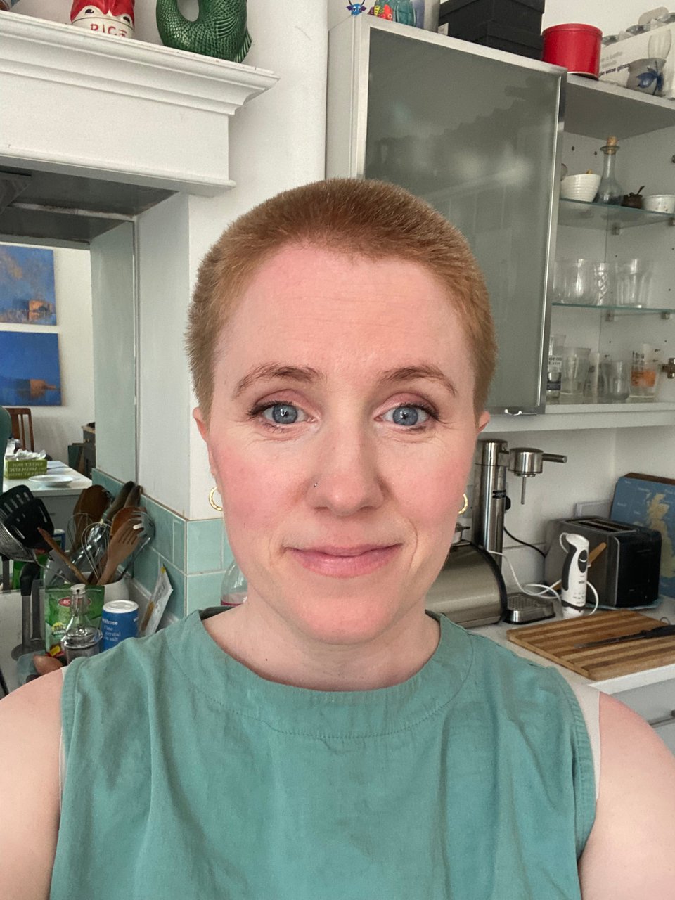 White woman, with shaved ginger hair, wearing a teal sleeveless top and smiling at the camera.
