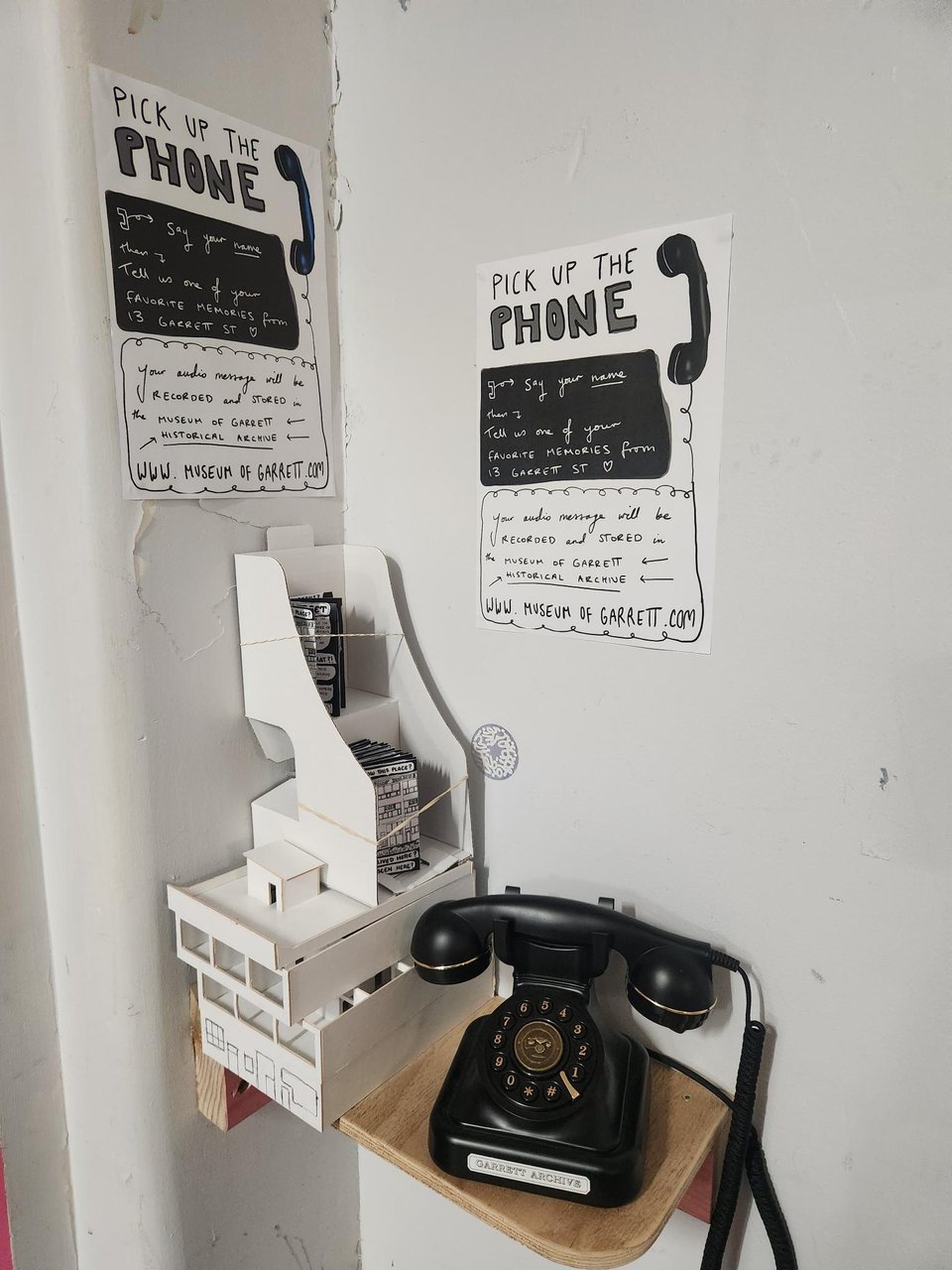 Image of black vintage-looking landline phone sitting on a small wooden shelf. A sign behind it reads 'PICK UP THE PHONE'