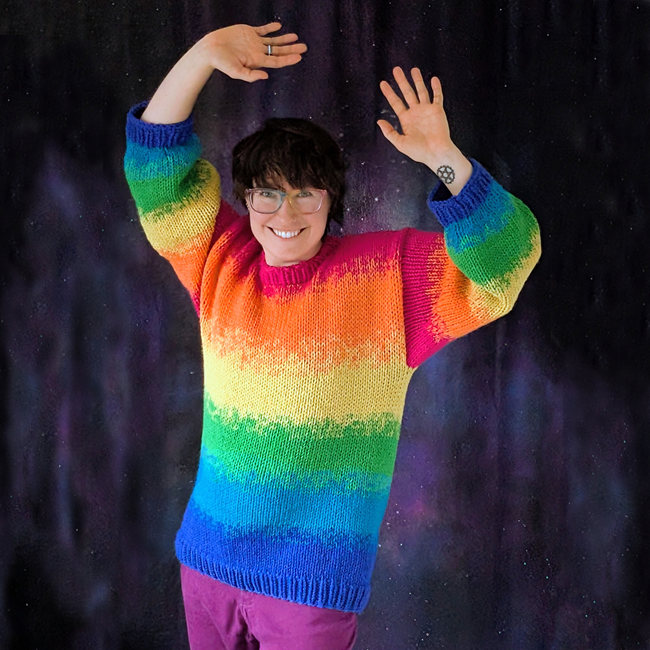 a photo of Maia from the waist up, hands in the air happily, wears a brightly colored, squishy oversized rainbow sweater.