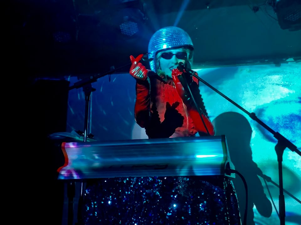 Photo of Ora the Molecule live at El Cid in Los Angeles. Performer dressed in a jumpsuit, gloves and disco ball helmet and is standing at a microphone behind a synthesizer while singing and snapping her fingers.