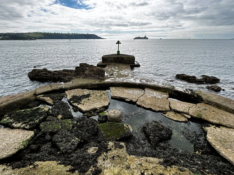Looking over Plymouth Sound, with a cruise ship anchored in it. In the foreground are the remains of a semi-circular concrete platform that has been broken up, and is slick with seaweed. At the tip of it is a short path to a triangular block toped with a green triangular navigational aid.